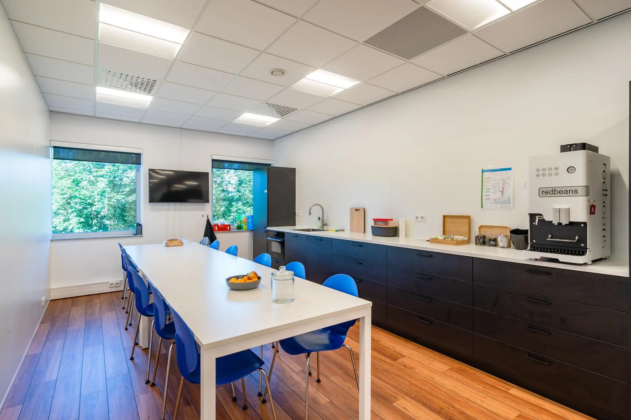 Moderne kantine met lange witte tafel, blauwe stoelen, koffieautomaat en uitzicht op groen bij Rivium Promenade.